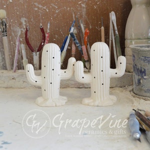 May include: Two white ceramic cactus sculptures with black dots and grooves. The cactus sculptures are on a white surface with tools and a bucket in the background.
