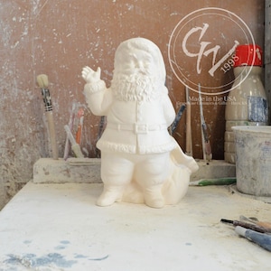 Puede incluir: Figura de cerámica blanca de Papá Noel con una bolsa de regalos. Está saludando con la mano izquierda. La figura está sin pintar y lista para ser decorada.