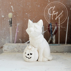 Peut inclure: Une figurine de chat en céramique blanche assise à côté d'une petite citrouille avec un visage sculpté. Le chat est assis droit et la citrouille est au sol. L'arrière-plan comprend des outils et le texte "Made in the USA".