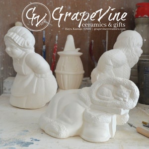 以下が含まれることがあります： 白いセラミック製の置物。女の子、ブーツの中で眠る猫、籠を持った人。置物は木製の台の上に置かれている。背景には「Grapevine ceramics & gifts」の文字が見える。