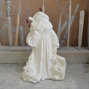 Puede incluir: Figura de cerámica blanca de Papá Noel sosteniendo una bolsa de regalos y un árbol de Navidad. La figura está de pie sobre una superficie gris.