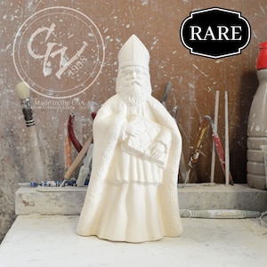 May include: A cream-colored ceramic statue of St. Nicholas holding a book, with a long white beard and a tall hat. The statue is labeled "RARE" and "Made in the USA" with the year "1998". The statue is on a work surface.