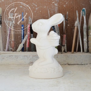 May include: White ceramic bunny figurine holding a basket of eggs. The bunny is standing on a round base with a decorative design. The figurine is made in the USA.