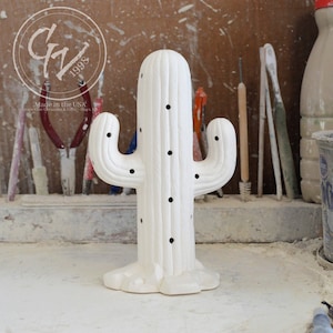 May include: A white ceramic cactus-shaped decorative item with black dot details. The cactus has two arms and a textured surface. The item is made in the USA, as indicated by the text on the image. The background includes tools and a workshop setting.