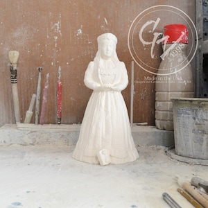 May include: White ceramic figurine of a girl wearing a crown and a long dress. The figurine is standing on a white surface. The background is a wooden wall with tools and a bottle of liquid.