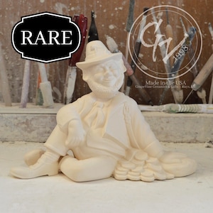 May include: A cream-colored ceramic leprechaun figurine, seated with a pile of coins. The figurine wears a hat and has a beard. The word "RARE" is in a black and white banner. The background includes tools.