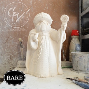 Puede incluir: Figura de cerámica blanca de Papá Noel con una túnica de piel y sosteniendo una copa. La figura está de pie sobre una mesa con un pincel y un cubo de pintura en el fondo. La palabra "RARE" está impresa en una etiqueta negra en primer plano.