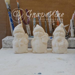 May include: Three unpainted ceramic snowman figurines. Each snowman wears a hat and scarf, and holds a different item: a snowflake, a lantern, and a wrapped gift. The background includes paintbrushes and the text "GrapeVine ceramics & gifts".