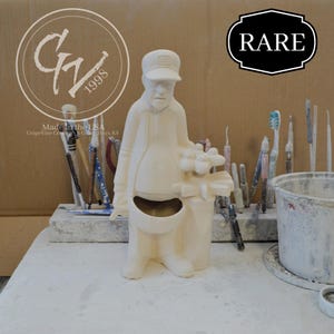May include: A cream-colored ceramic figurine of a man in a cap and coat, holding a bouquet. The figurine has a pocket-like opening in the front. The word "RARE" is in a black and white banner. The image also includes the text "Made in the USA" and "GrapeVine Ceramics & Gifts, Hays, KS."
