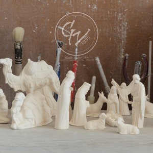 May include: A set of unpainted ceramic figurines depicting the Nativity scene, including Mary, Joseph, baby Jesus, a donkey, a camel, and two shepherds. The figurines are arranged on a table with a wooden background. Made in the USA.