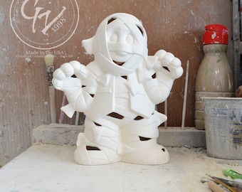 May include: White ceramic mummy figurine with a cutout design. The figurine is standing on a table with a paintbrush and other craft supplies.