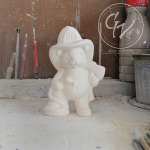 May include: White ceramic figurine of a bear wearing a firefighter hat and holding an axe. The bear is standing on a white surface.