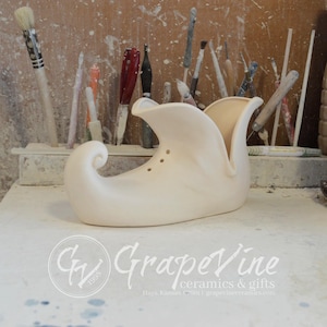 Puede incluir: Una maceta de cerámica con forma de zapato de elfo en color crema. El zapato tiene una punta rizada y una abertura acampanada. Se ven tres pequeños agujeros en el lateral. El logotipo de GrapeVine ceramics & gifts está en la parte inferior.