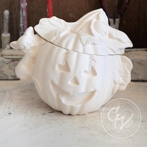 Op de afbeelding: Witte keramische pompoen met een deksel in de vorm van een vleermuis. De pompoen heeft een gesneden jack-o'-lantern gezicht en de vleermuis heeft een kleine kat op zijn rug. Gemaakt in de VS.