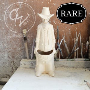 May include: White ceramic cowboy figurine wearing a hat and holding a pot. The figurine is labeled "RARE" and "Made in the USA".