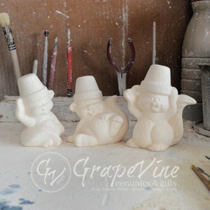 May include: Three white ceramic figurines of squirrels wearing hats. The squirrels are in different poses, one is sitting, one is lying down, and one is standing. The figurines are on a wooden surface.