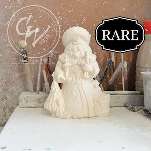 May include: A cream-colored Santa Claus figurine with a round hat and holding objects. The figurine has intricate details and is labeled "RARE" in a black frame. The background includes paintbrushes and other art supplies.