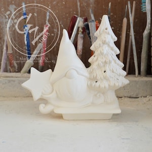 Może przedstawiać: Biała ceramiczna figurka gnoma ciągnącego sanie z choinką. Gnom ma gwiazdę na czapce i brodę. Wyprodukowano w USA. Grapevine Ceramics & Gifts, Hays, KS.