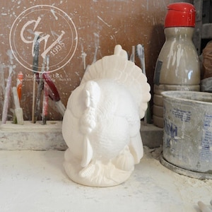 May include: White ceramic turkey figurine with a detailed tail and feathers. The figurine is standing on a base with leaves and acorns. Made in the USA.