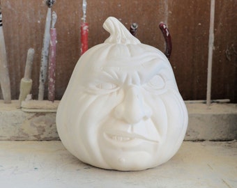 May include: White ceramic pumpkin with a detailed, expressive face. The pumpkin has a wide, toothy grin and a furrowed brow, giving it a mischievous and slightly menacing look.