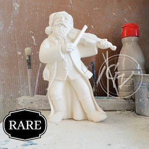 Peut inclure: Figurines en céramique blanche de Père Noël jouant du violon. La figurine est debout sur une surface en bois avec des pinceaux et d'autres fournitures d'artisanat en arrière-plan. La figurine est marquée "RARE" au premier plan.