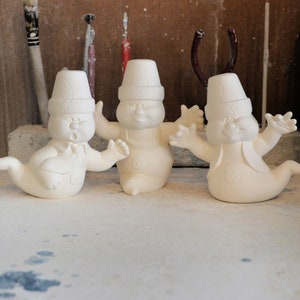 May include: Three white ceramic ghost figurines with a whimsical design. Each ghost has a hat and a friendly face. The ghosts are standing on a white surface.