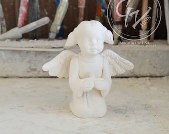 Może przedstawiać: Biała ceramiczna figurka klęczącego anioła z skrzydłami. Anioł ma słodki wyraz twarzy i trzyma ręce złożone do modlitwy. Figurka została wykonana w USA przez Grapevine Ceramics & Gifts.