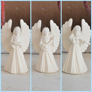 Puede incluir: Tres figuras de ángeles de cerámica blanca. Los ángeles llevan todos túnicas y tienen alas. Un ángel sostiene un libro, otro un instrumento musical y el último un violín.