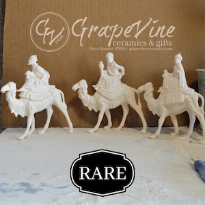 Puede incluir: Tres figuras de cerámica blanca de los Reyes Magos montados en camellos. Las figuras son detalladas y se encuentran sobre una superficie blanca. La palabra "RARE" está en un óvalo blanco y negro. El fondo tiene el texto "GrapeVine ceramics & gifts".