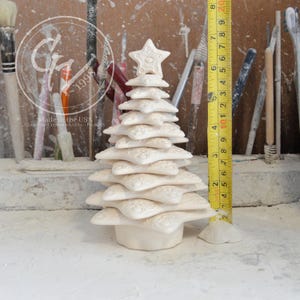 May include: A white ceramic Christmas tree ornament with a star on top. The tree is made of stacked stars with a textured surface. The ornament is approximately 7 inches tall.