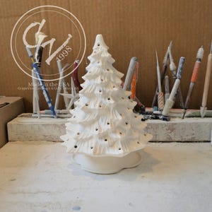 Op de afbeelding: Een wit keramieken kerstboombeeldje met een getextureerd, gelaagd takontwerp en talloze kleine ronde openingen voor lichtjes. Het stuk heeft een gladde, geschulpte voet en is uitgevoerd in een ongeverfde, biscuit-gebakken afwerking.