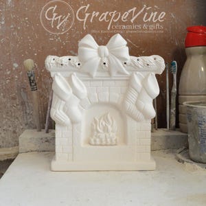 May include: White ceramic fireplace with a bow on top and two stockings hanging on the sides. The fireplace has a brick design and a fire in the hearth.