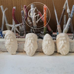 May include: Four unpainted ceramic Santa Claus face ornaments arranged in a row. Each piece features detailed facial hair and a hat. Behind the ornaments, various sculpting and pottery tools are standing upright in a wooden holder.