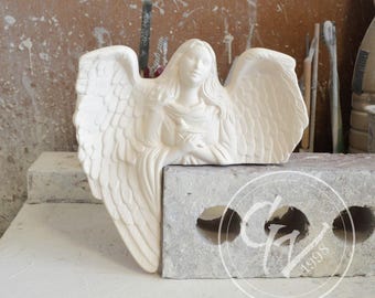 Może przedstawiać: Biała ceramiczna figurka anioła z rozłożonymi skrzydłami. Anioł trzyma gołębia w dłoniach. Figurka stoi na szarym cegle.