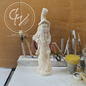 May include: A cream-colored ceramic wizard figurine with a long beard, hat, and staff. The figurine is standing on a white surface, with various art tools in the background. The image includes the text "Made in the USA" and "Grapevine Ceramics & Gifts, Hays, KS".