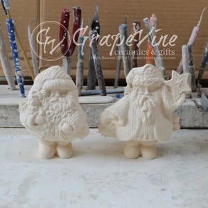 May include: Two unpainted ceramic Santa Claus figurines. One Santa holds a star, the other holds a small ornament. The figurines have detailed textures and are in a workshop setting. The text "GrapeVine ceramics & gifts" is visible.
