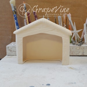 Può includere: Una stalla per presepe in ceramica color crema con tetto strutturato e apertura ad arco. Il logo GrapeVine Ceramics & Gifts è visibile sullo sfondo, insieme a pennelli e altri materiali artistici. La stalla è un oggetto fatto a mano.