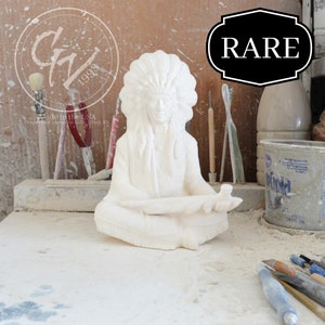 Puede incluir: Figurilla de cerámica blanca de un hombre nativo americano con tocado y pipa. La figurilla está sentada en una mesa con herramientas y otros objetos en el fondo. La palabra "RARE" está escrita en un letrero negro sobre la figurilla.