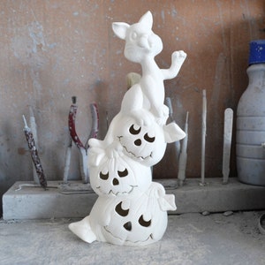 Puede incluir: Decoración de Halloween de cerámica blanca con un gato encaramado en la cima de una pila de tres calabazas. Las calabazas tienen caras talladas con ojos, narices y bocas.