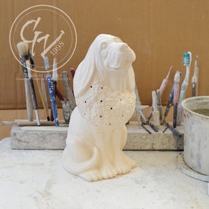 May include: A bisque ceramic dog figurine, featuring a seated pose with detailed fur and a textured chest. The dog has long, floppy ears and a gentle expression. The image includes the text "Made in the USA" and "Grapevine Ceramics & Haycks, KS".