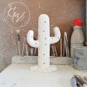 Puede incluir: Escultura de cactus de cerámica blanca con una superficie texturizada y pequeños agujeros. El cactus está de pie sobre una mesa blanca con un estante de madera en el fondo.