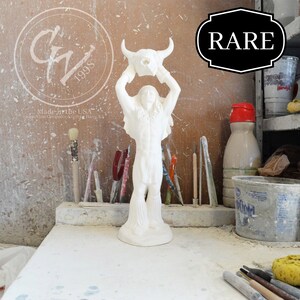Op de afbeelding: Witte keramische figuur van een indiaanse man die een buffel schedel boven zijn hoofd houdt. De figuur staat op een witte tafel met kwasten en andere gereedschappen op de achtergrond. Het woord "RARE" staat in het zwart op een witte achtergrond in de rechterbovenhoek van de afbeelding.