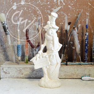 Puede incluir: Figura de muñeco de nieve de cerámica blanca con un cartel que dice "MERRY CHRISTMAS". El muñeco de nieve lleva un sombrero de copa y una bufanda. El fondo incluye herramientas de arte y el texto "Made in the USA."