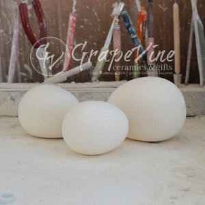 May include: Three white ceramic eggs of varying sizes. The eggs are sitting on a light gray surface. The background is a wooden shelf with various tools and a sign that reads "Grapevine ceramics & gifts Hays, Kansas 67601 | grapevineceramics.com"
