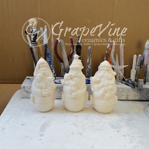 May include: Three unpainted ceramic Santa Claus figurines. Each figure has a unique design, with details like beards, hats, and decorative elements. The figurines are on a white surface, with tools and the GrapeVine Ceramics & Gifts logo in the background.