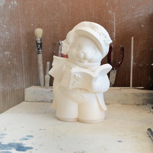 Könnte beinhalten: Eine unbemalte Biskuit-Keramikfigur eines Bären mit Hut und Schal, der ein Buch hält. Der Bär trägt Winterkleidung und liest ein Buch mit einem dekorativen Design. Die Figur steht auf einer weißen Oberfläche.