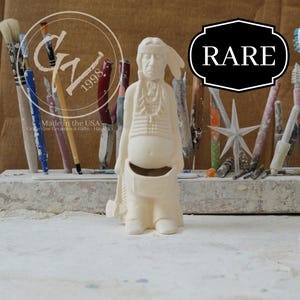May include: A cream-colored ceramic Native American figure with a headdress, holding a tomahawk. The figure has a detailed vest and a pouch. The word "RARE" is in a black and white label. The background includes paint brushes and tools.