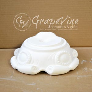 Könnte beinhalten: Ein weißes dekoratives Keramikstück mit einer einzigartigen, abgerundeten Form. Das Stück weist geprägte, blütenartige Designs und einen erhöhten Mittelbereich auf. Das Logo von GrapeVine Ceramics & Gifts ist im Hintergrund sichtbar.