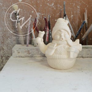 May include: A ceramic snowman figurine, unpainted, with a hat, scarf, and mittens. The snowman holds a lantern and a bell. The base of the snowman is a rounded shape. The text "Made in the USA" is visible.