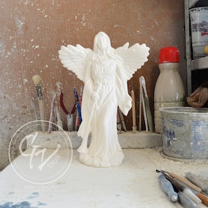 Op de afbeelding: Witte keramische engelfiguur met uitgespreide vleugels, die een trompet in de ene hand vasthoudt. De engel staat op een voetstuk en draagt een lange, vloeiende robe. Het beeldje staat in een werkplaats met gereedschap en kwasten op de achtergrond.